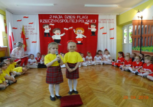 Dwie dziewczynki w żółtych podkoszukach i czerwonych spódniczkach w kratę recytują wiersz.